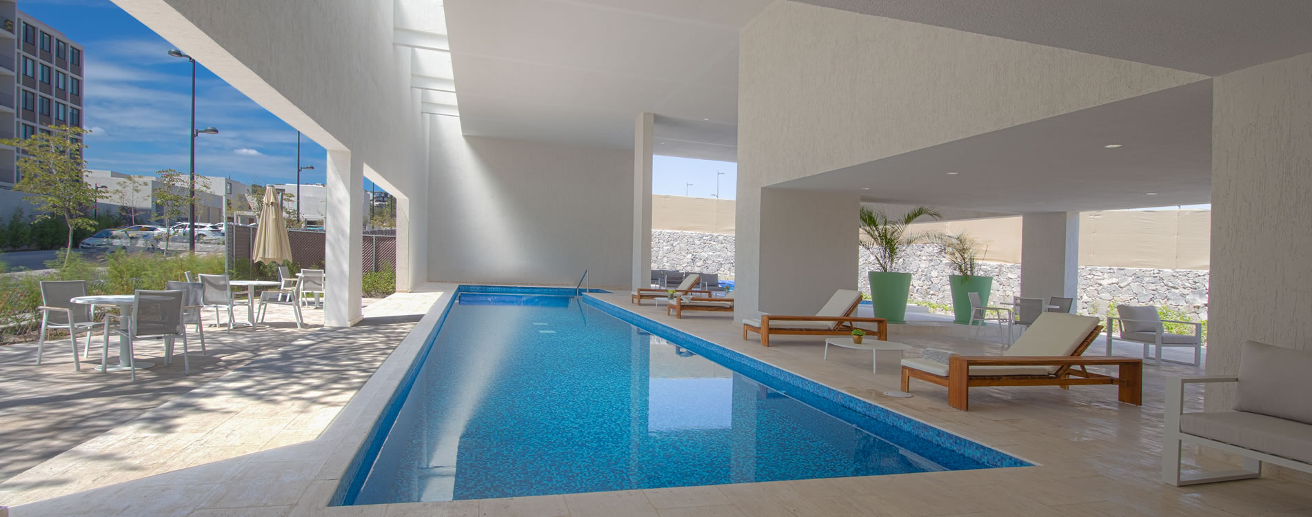 Alberca interior con azulejos azules y área de descanso con sillas y mesas blancas.